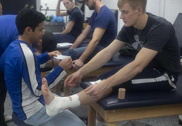 Two male Graceland students practicing ankle wrapping in the athletic training room.
