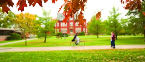 Autumn leaves frame a cyclist and pedestrian on a campus path, red brick building in background.