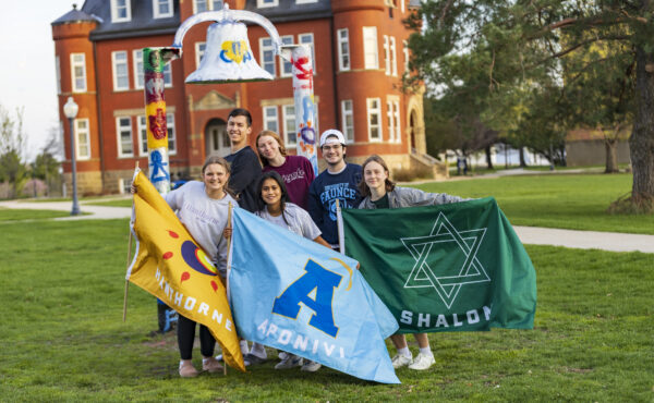 A group of college students holding flags standing on grass in front of a painted bell