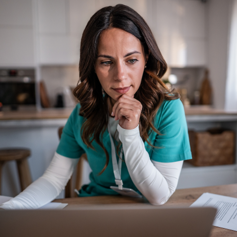 Woman in a teal scrub top working intently on a laptop in a bright kitchen setting, papers scattered on the table